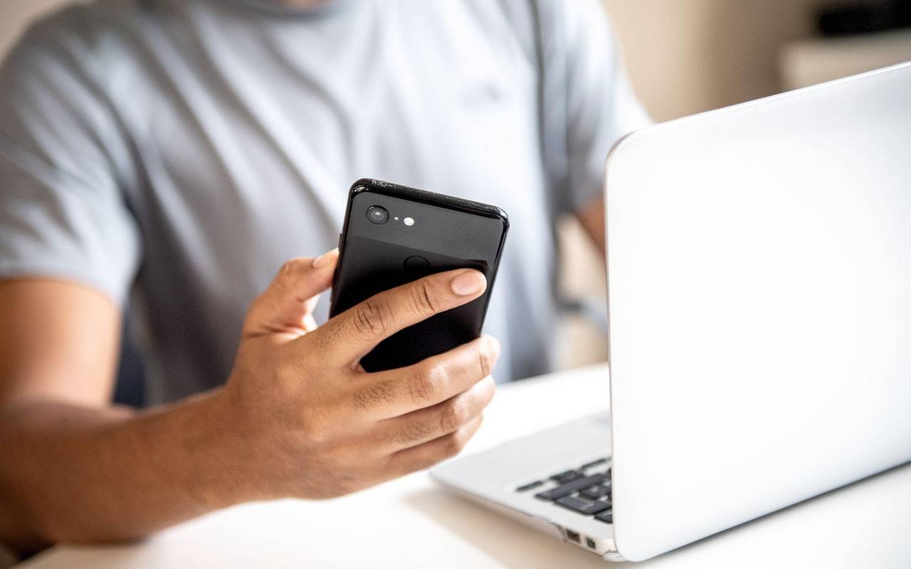 Mann hält vor einem Laptop sein Smartphone in der Hand