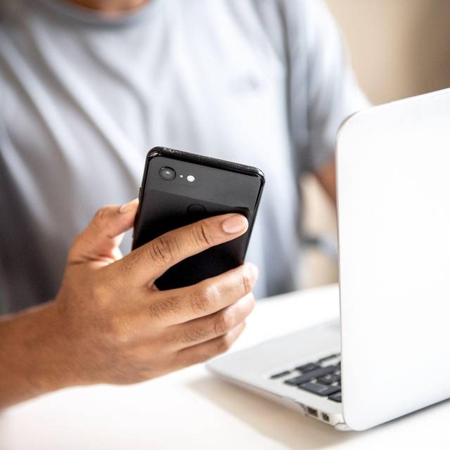 Mann hält vor einem Laptop sein Smartphone in der Hand